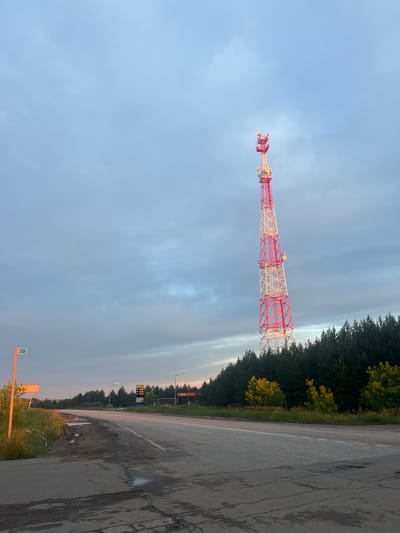 The image depicts a landscape scene near the city of Golyan, Russia. In the foreground, a paved road with cracked asphalt and puddles stretches into the distance. The road is bordered by sparse green grass and some vegetation. To the left of the road, a tall pole with a green and orange sign is visible. Further down the road, a gas station sign with prices displayed and a building with red signage are present. A dense line of dark green evergreen trees forms the backdrop behind the road and gas station. Dominating the right side of the image is a tall, red and white lattice telecommunications tower, reaching high into the overcast sky. The sky is filled with gray and light blue clouds, with some hints of pink or orange near the horizon, suggesting it might be dawn or dusk. The overall scene conveys a sense of a rural or roadside environment in Russia.