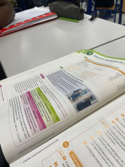 This image depicts an interior scene, specifically a classroom or study area, located in Hérouville-Saint-Clair, France. The primary subject is an open textbook, written in French and published by NATHAN, resting on a clean, white desk surface. The visible pages display content related to business activity, with the heading "4 Mesurer l'activité de l'entreprise" (Measuring business activity). Sections include tables of numerical data and categorized information such as "Indicateurs de productivité" (Productivity indicators) and "Indicateurs sociaux" (Social indicators). A small color photograph within the textbook illustrates two individuals: a woman with glasses looking at a monitor, and a man in a suit examining documents. To the upper left of the textbook, a red binder with white polka dots is partially visible. A dark-skinned human arm rests on the desk surface near this binder. Further to the left and rear of the binder, a dark gray pencil case and a green pen are present on the desk. The background is blurred, showing blue-framed classroom furniture consistent with additional desks and chairs.