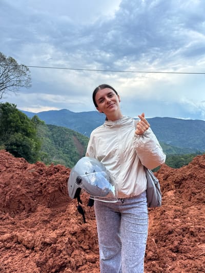 A young woman stands outdoors in a hilly, rural landscape, holding a gray motorcycle helmet in her left arm. She is wearing a light-colored zippered jacket and light-wash jeans, and has her right hand raised, forming a heart shape with her thumb and index finger. Behind her, the terrain is a steep, reddish-brown slope of loose soil, likely the result of a landslide. In the background, lush green mountains rise under a cloudy sky. The location is described as Nao Sẻ, Vietnam, suggesting a remote, possibly mountainous region.