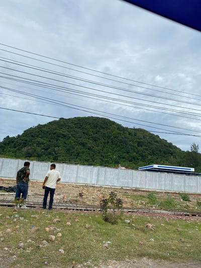 The image, taken near Lamkruet, Indonesia, shows two men standing near a construction site. A green, densely forested hill rises in the background. Overhead power lines are visible on the left side of the frame against a cloudy sky. A long, blue and white structure sits atop a concrete wall near the hill. The construction site is adjacent to a grassy area littered with rocks and debris. One man is wearing a white shirt and blue jeans, and the other wears a camouflage shirt and blue jeans.