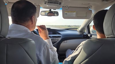 Two individuals are seated in the front seats of a car, facing forward and driving on a paved road. The person in the driver's seat, viewed from behind, is wearing a white collared shirt and has dark hair. Their right hand is raised and holding a light-colored object, possibly a toothpick, near their mouth. The person in the passenger seat, also viewed from behind, has dark hair and is wearing a light-colored garment.

The car's interior is visible, including the dashboard, steering wheel, rearview mirror, and sun visors. The rearview mirror reflects the face of the driver. The sun visors have warning labels attached. The car is in motion, indicated by the road ahead and the landscape passing by.

The environment outside the car appears to be arid and flat, with sparse vegetation and a clear sky. The scene suggests a journey on a highway in a potentially remote or rural area. The location is identified as Sakarçäge District, Turkmenistan.
