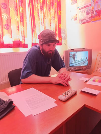 A man wearing a dark blue t-shirt and a bucket hat sits at a wooden desk in what appears to be a small room. He has a beard and his hands are clasped in front of him. On the desk, there is a small, old television set displaying a black and white image, a remote control, papers, envelopes, and a pen. In the background, red curtains with a patterned design hang over a radiator. Posters are visible on the wall behind the man. The lighting in the room has a strong red hue. This scene is located in the city of Ladyzhyn, Ukraine.