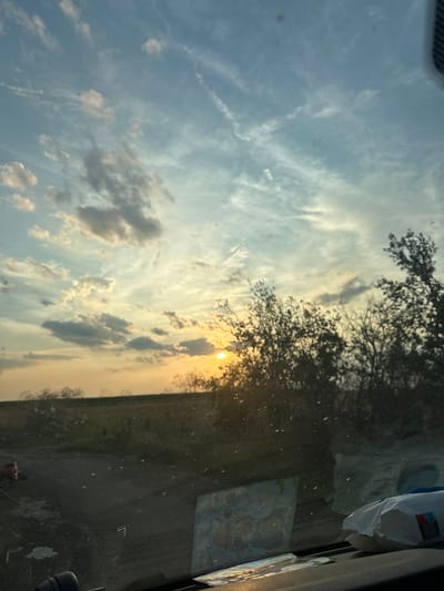 The image captures a serene sunset over a rural landscape, likely in the vicinity of Mlekarevo, Bulgaria. The sky is a canvas of soft blues and yellows, with wispy clouds stretching across, illuminated by the setting sun peeking through the horizon. In the foreground, silhouetted trees and bushes create a natural frame, their dark forms contrasting with the vibrant sky. The ground appears to be a dirt road or path, with patches of dry grass and vegetation suggesting a dry, warm climate. On the left side of the image, a small red object with wheels, possibly a toy or a piece of equipment, is partially visible. The scene is viewed from inside a vehicle, as evidenced by the windshield's reflection of dust and water droplets, and the presence of items on the dashboard, including a packet of wet wipes and a decorative item with cartoon characters. The overall impression is one of peace and tranquility, a moment captured as day transitions into night in a natural, undeveloped setting.