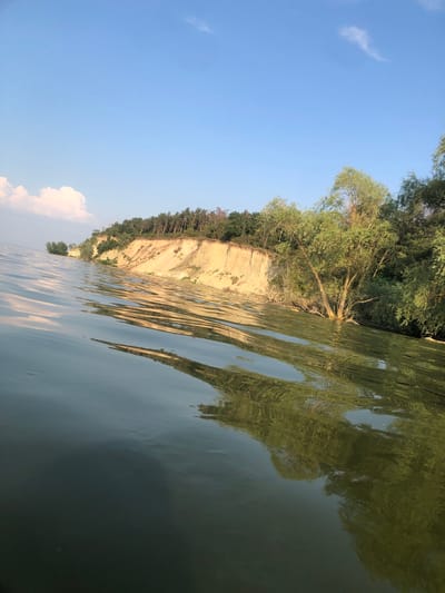 The image depicts a body of water with a steep, sandy cliff face on the right side. The cliff is covered with trees and vegetation at the top. The water in the foreground is rippled and reflects the trees and sky. The sky is blue with a few scattered clouds. The scene appears to be a natural landscape, possibly a riverbank or shoreline. The specific location in Nahirne, Ukraine, is not visually identifiable from the image alone, but the natural elements suggest a rural or semi-rural setting. There are no people or man-made structures immediately visible.