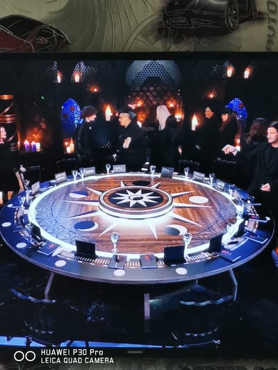 The image shows a dimly lit room with a large, round table in the center. The table is decorated with a compass rose design and has several place settings with glasses and what appear to be notebooks or books. Several people are seated or standing around the table, all dressed in dark clothing. The atmosphere appears to be formal or ceremonial. The background shows ornate lamps and stained-glass windows. The bottom of the image displays a "HUAWEI P30 Pro LEICA QUAD CAMERA" watermark and a "SAMSUNG" logo. The overall scene does not provide any specific clues to determine its location within the city of Bad Oeynhausen, Germany.