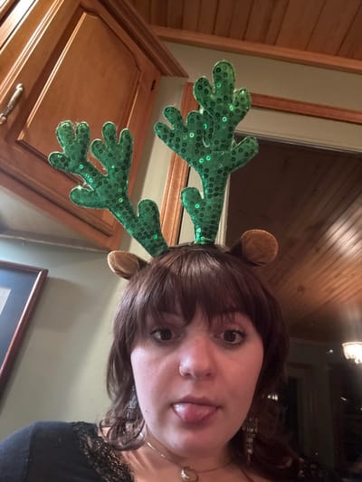 A person wearing reindeer antlers, showing her tongue. The antlers are green with sequins and have brown ears. The background includes wooden cabinets, a window, and a framed picture. The person has brown hair with bangs and is wearing a black shirt with lace.
