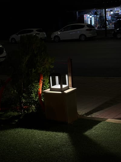 The image depicts a nighttime street scene in Saâda, Morocco, characterized by artificial lighting and shadows. The primary subject is a decorative wooden structure, possibly a stylized well or lantern holder, illuminated from within. Its light casts strong shadows on the surrounding elements.

To the right of the wooden structure, artificial green turf covers the ground, bordered by a brick-patterned pathway or wall section. A dark bush or small tree with green foliage is situated near the wooden structure, with a bright orange corrugated pipe or rope wrapping around its base and extending into the foreground.

In the background, on the left, a brightly lit shop is visible, displaying various items on shelves through its large windows, suggesting a retail business. Two white cars are parked on what appears to be a paved street or parking area in front of the shop. Another partial view of a car is visible further down the street to the right.

The overall scene is dark, with light sources primarily from the shop, the interior of the wooden structure, and potentially car headlights or other street lighting, creating a high-contrast image. No people or specific activities are visible. No discernible text is present in the image.