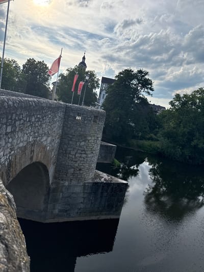 The image captures a view of the Lahn river in Wetzlar, Germany. A stone bridge, likely historic with its rough-hewn blocks, dominates the foreground. One arch of the bridge is visible, leading into a dark, still body of water that reflects the sky and surrounding trees. The riverbank is lush with green trees, some of which cast shadows on the water.

In the background, a building with a distinct dome and steeple suggests a church or cathedral. Red flags with white lettering are displayed on poles along the riverbank, adding a touch of color to the scene. The sky is partly cloudy, with dramatic, wispy clouds illuminated by the setting sun, casting a warm glow on the left side of the image. The overall mood is tranquil and picturesque, hinting at a late afternoon or early evening. There is no visible text on the flags that can be clearly deciphered in the provided image.