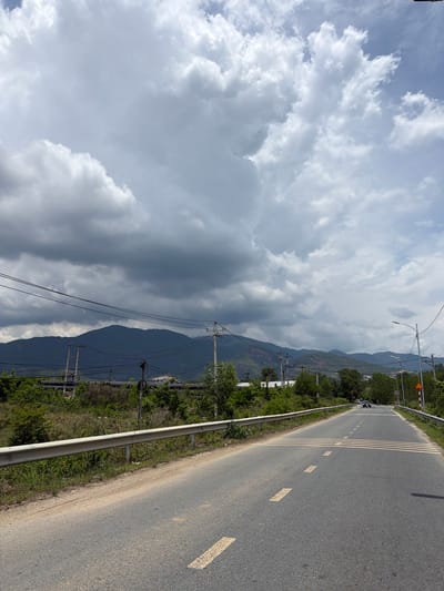 The image shows a paved road in the city of Dien Tho, Vietnam, stretching into the distance towards a range of mountains. The sky is partly cloudy with dramatic, dark gray clouds dominating the upper portion of the frame, contrasting with lighter, puffy white clouds below. The road has yellow dashed lane markings and a pedestrian crossing marked with yellow stripes. A metal guardrail runs along the left side of the road, separating it from overgrown vegetation. Several utility poles with electrical wires are visible along the roadside and in the middle ground, with some industrial-looking pipes also present. In the distance, a few trees and buildings can be seen at the base of the mountains. A car is visible on the road in the distance, and there appears to be a motorbike behind it. The overall impression is of a somewhat industrial or rural landscape under a brewing storm.