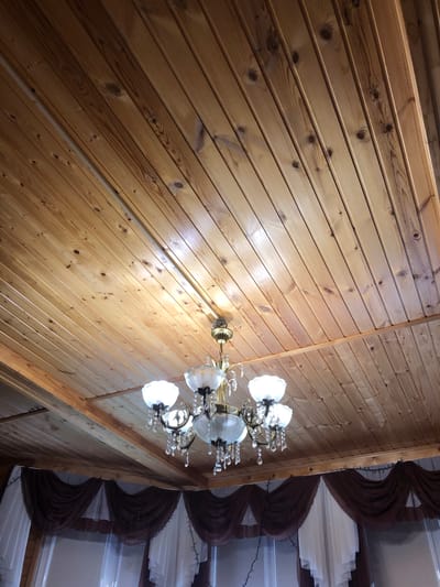 The image shows a wooden plank ceiling with a chandelier. The chandelier has a brass-colored frame and multiple glass shades, adorned with crystal droplets. The ceiling is made of light-colored wood planks, oriented diagonally from the bottom left to the top right of the frame. The room below the ceiling is not fully visible but includes draped brown and white curtains. The lighting in the image suggests it was taken indoors, likely in a residential setting. The specific location of Московский Ветеран Снт, Russia, is not discernible from the visual content of the image alone.