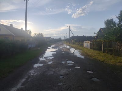 This image captures a rural scene in the Yaglevitski village council, Belarus, featuring a dirt road lined with houses and trees. The road is partially covered in puddles reflecting the cloudy sky, suggesting recent rainfall. Power lines and poles are visible, characteristic of rural infrastructure. The houses appear to be of traditional design, with wooden fences and small yards. The overall impression is of a quiet, serene village setting under a partly sunny sky.