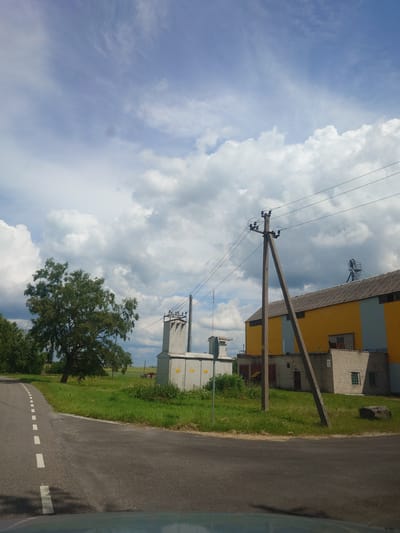 The image shows a rural scene in Alavere, Estonia, on a cloudy day. A paved road with a dashed white line curves into the distance on the right side of the frame. To the left of the road is a grassy area with several large trees, a power transformer, and electrical poles with wires running overhead. Further back, a yellow and grey building with a corrugated roof stands behind a smaller brick building. The sky is filled with dramatic, fluffy white clouds against a light blue background, suggesting it may be mid-day. The overall impression is one of a quiet, functional rural landscape with visible infrastructure. No people are visible in the image, and no text is discernible.