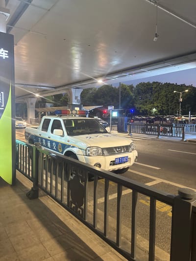 A white pickup truck with flashing red and blue lights on its roof is parked on a street at night in Xinwu District, China. The truck has blue and white markings on its side that appear to be official insignia. The license plate reads "B 71UE5". The truck is behind a black metal fence. In the background, there are trees, streetlights, other vehicles, and an elevated structure. The overall lighting suggests it is dusk or early evening.