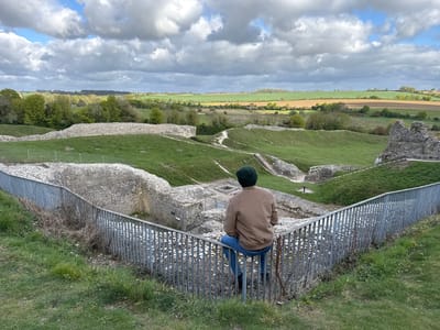 A lone figure, viewed from behind, sits atop a metal fence, observing a historical landscape under a partly cloudy sky. The man wears a dark green beanie, a brown bomber jacket, blue jeans, and dark boots. He is perched on a sturdy grey metal fence composed of vertical bars, with a walking stick or cane resting beside him.
The setting is an archaeological site or ancient ruins in King's Lynn and West Norfolk, United Kingdom. Below the fence and the viewer, extensive stone foundations and low walls of an old structure are visible, partially covered by green grass. Dirt paths wind through these ruins and ascend grassy mounds. In the mid-ground, a lush valley unfolds, dotted with various green trees and vegetation.
Beyond the historical site, the rural landscape stretches to the horizon, featuring a mosaic of cultivated fields in shades of green and brown. A line of trees forms the distant edge of the land. The sky is filled with large, dramatic grey clouds, though patches of blue sky are also visible, suggesting a dynamic weather pattern typical of daytime. The overall atmosphere is one of quiet contemplation, with the man seemingly absorbed in the panoramic view of the ancient ruins and the expansive countryside. Small informational signs are scattered within the archaeological dig, but their text is unreadable from this distance.