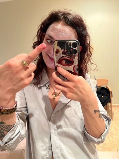 A person is indoors taking a selfie with a phone that has a case decorated with stylized eyes. The person's face appears to have a mask or cream applied. The person is wearing a light gray button-down shirt, jewelry including a ring, a bracelet, and a necklace. The background includes a beige wall and a shelf with some items on it. The image does not contain sufficient information to verify the location in the city of Puçol, Spain.