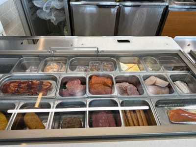 This media file shows a close-up, eye-level view of a refrigerated food display case, likely in a restaurant or deli. The display case is filled with various food items organized in separate compartments. The items include different types of meats, such as sliced salami, ham, and what appears to be a breaded chicken patty. There are also some cheese slices and other unidentifiable food items. Some compartments have small handwritten labels with dates and times, for example, "18.06 10:00" and "10.06 10:00". In the background, stainless steel appliances and white plastic bags are visible, suggesting a commercial kitchen setting. The overall lighting is bright, indicating an indoor environment, possibly during daytime. There are no people visible in the frame. The scene depicts a well-stocked food display, ready for service. The location context is Mikhailovka, Russia.
