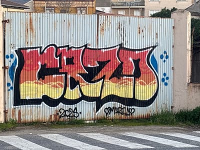 The image depicts a corrugated metal gate in Piedrahíta, Spain, featuring prominent graffiti art. The gate, appearing weathered and light blue, exhibits significant rust discoloration, particularly along its lower edge and seams. It consists of two panels, hinged on the left. A large, stylized graffiti piece spans the gate, rendered with black outlines, a white inner border, and a fill gradient from red at the top, transitioning to orange in the middle, and yellow at the bottom. The letter fills incorporate wavy, cloud-like patterns. Blue droplet shapes and dots accent the far left and right edges of the artwork. Below the main graffiti, "2023." is tagged on the left, and "♡MARTIAS♡" is tagged on the right. In the foreground, a narrow strip of dirt with sparse green vegetation separates the gate from a paved road. The road surface displays white diagonal stripes, indicative of a pedestrian crossing. Behind the gate, multi-story buildings, possibly residential, are partially visible along with some bare tree branches. No individuals or active elements are present in the scene.
