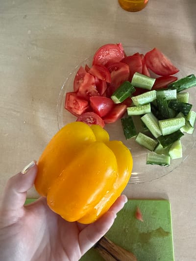 In this overhead shot, a hand holds a bright yellow bell pepper, its skin glistening with water droplets. The pepper is positioned in the foreground, partially obscuring a clear glass plate filled with chopped vegetables. Sliced red tomatoes and cut green cucumbers are arranged on the plate, appearing to be prepared for a salad.
To the right of the plate, a vibrant green cutting board is visible, also wet, suggesting recent food preparation. A wooden utensil rests on the cutting board. The background is a textured surface, likely a table, in a neutral, light brown tone.
The lighting is bright and natural, casting soft shadows, and there are no discernible time of day or weather cues. The setting appears to be a kitchen or dining area. The focus is on the fresh ingredients and the process of preparing food. No text is visible in the image. The location context provided indicates this scene is in Nizhne-Chufichevo, Russia.