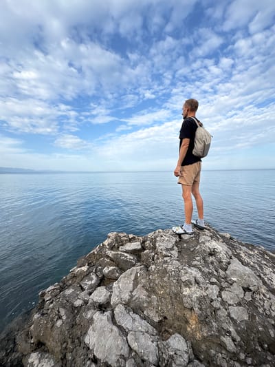 The image shows a man standing on the edge of a rocky cliff overlooking a body of water. The sky is visible in the background, featuring blue sky with scattered clouds. The man is wearing a black shirt, tan shorts, a backpack, and white socks with slides. The cliff appears to be composed of grey rock formations. It is not possible to verify the location is in Bondarenkovo, Ukraine from this image.
