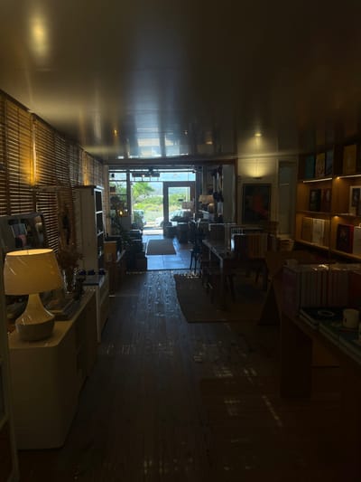 The image shows a long, narrow room with light streaming in from a door at the far end, revealing a view of greenery. Bookshelves line the upper walls of the room, filled with books. Several tables and chairs are situated in the middle of the room. The floor is dark wood and patterned. A lamp and furniture are visible in the foreground. The room seems to be either a bookstore or a library.