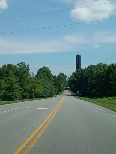 This is a photograph of a road curving through a wooded area on a clear day. The camera is oriented vertically, and the scene is viewed from a slightly elevated perspective. A tall, dark structure, possibly a water tower or communication tower, stands prominently against the bright blue sky on the left side of the image. The tower has a series of platforms or levels visible. Power lines stretch across the sky.

The road itself is asphalt and features a white line on the left edge and double yellow lines in the center. A white arrow indicating a turn is painted on the road surface. A few cars are visible on the road in the distance. The right side of the road is bordered by a well-maintained grassy area, transitioning into a dense line of green trees. The trees are lush and full, suggesting a warm season, likely summer.

The lighting indicates it is daytime, with clear skies and visible clouds. The overall atmosphere is serene and natural. No people are clearly identifiable in the image. The location is identified as Versailles, United States. The image appears to be taken from a moving vehicle, given the perspective and the slight blur in some areas.