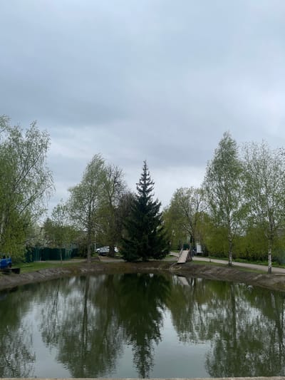 The image depicts a body of water, possibly a pond or canal, located in what appears to be a park or recreational area. The scene is characterized by trees lining the water's edge, their reflections visible in the water. The sky is overcast, suggesting a cloudy day. No people are clearly visible in the image. The location is specified as Хлябово, Russia.
