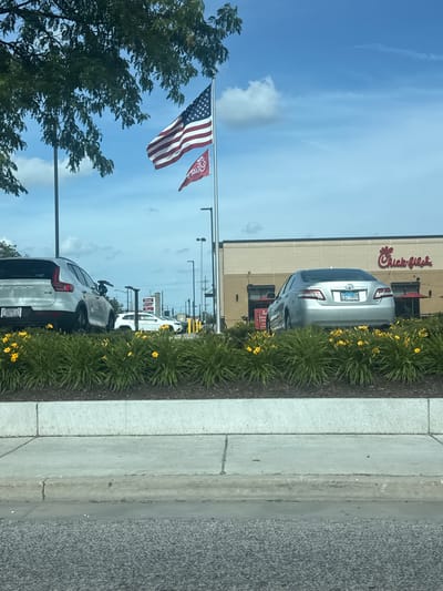 In Niles, United States of America, a sunny day unfolds with a Chick-fil-A restaurant as the backdrop. The American flag flies proudly from a tall pole, accompanied by a smaller maroon flag featuring the Chick-fil-A logo. In the foreground, a silver Volvo SUV and a silver Toyota Camry are parked. The cars are positioned amidst lush green bushes dotted with vibrant yellow flowers, suggesting a well-maintained landscape. The restaurant building, with its distinctive brick facade and large windows, stands behind the vehicles. Outdoor umbrellas are visible outside the restaurant. The sky is a bright blue with scattered white clouds, and tall streetlights punctuate the scene. The asphalt road in the very bottom foreground indicates this is likely a street view.