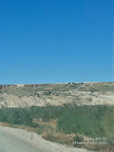 The image shows a barren landscape under a clear blue sky. In the foreground, a dirt road runs alongside a dense thicket of green vegetation. Beyond the vegetation, a light-colored, rocky hill rises, carved with roads and dotted with sparse green plants. The top of the hill features more developed structures, including what appear to be a road network, some buildings, and possibly some industrial installations. The overall scene depicts a somewhat arid and undeveloped area. A text overlay in the bottom right corner indicates "Galaxy A56 5G 23 июня 2026 г. 10:15", suggesting the image was taken on June 23, 2026, at 10:15 AM. The location is identified as Unknown, Uzbekistan.