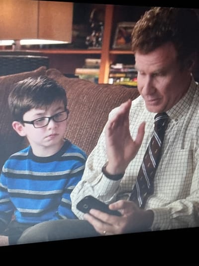 The image shows a close-up of two people sitting on a couch. On the left is a young boy wearing black-rimmed glasses and a blue and gray striped long-sleeved shirt. He is looking downwards. To the right of the boy is a man with reddish-brown hair wearing a light-colored checkered button-down shirt and a striped tie. His right hand is raised in a gesturing motion, and his left hand is holding a black smartphone. The background appears to be an indoor setting with a bookshelf visible behind the man. The scene does not contain any information to suggest a location in Enevo, Bulgaria.