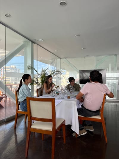 Four people are seated at a table in a restaurant with large glass windows overlooking a city and ocean. The table is covered with a white tablecloth and set with place settings, including plates, glasses, and cutlery. A large potted plant is positioned behind the table. One person, a woman with dark hair pulled back in a ponytail, is wearing a light blue long-sleeved shirt and blue jeans. Next to her, a woman with long brown hair is wearing a white tank top. Across the table, a man with dark hair and glasses is wearing a green shirt. To his right, a man with dark hair is wearing a pink t-shirt and black jeans. The floor is dark wood, and the ceiling has recessed lighting. The backdrop outside the window shows buildings and a bright sky, suggesting a coastal city environment. The restaurant appears to be part of a yacht club, as indicated by text on a screen in the background.