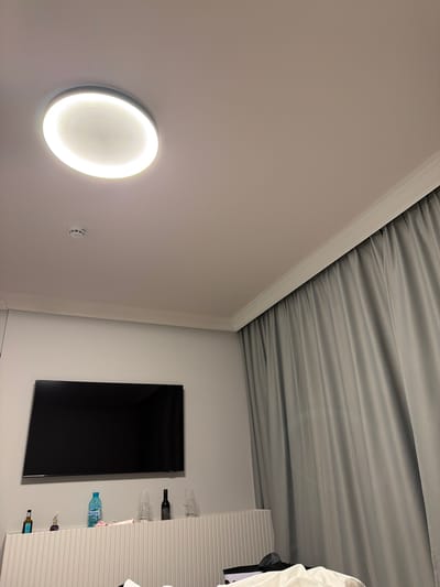 The image displays the interior of a modern room, likely a hotel or guest room, with a distinctive design. The perspective is slightly upwards, showing white walls and a unique gray, pleated fabric ceiling that creates a soft, draped texture. A white molding separates the walls from the ceiling.
On the left wall, a prominent circular light fixture emits a bright, warm glow, illuminating the immediate area. Below this light, a small, white, circular device with three horizontal slots is mounted, possibly a thermostat, sensor, or alarm.
The right side of the room features a large, rectangular black screen, likely a wall-mounted television. Adjacent to the television, a tall, white cabinet or shelving unit with vertical ribbing holds several items. On its upper shelves, multiple empty wine glasses are stored upside down, alongside a dark bottle that appears to be wine. A blue plastic water bottle is visible on a lower shelf, with text on its label that is difficult to read but could be a brand name like "DEVIN." Below it, two smaller bottles, one with a purple top, are also displayed. A white piece of fabric, possibly a garment or towel, hangs over the far right side of the cabinet.
The room is illuminated by artificial light, indicating an indoor setting where natural light is not prominent. The overall aesthetic is clean and contemporary. The location is Нареченски бани, Bulgaria.