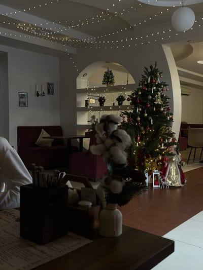 The scene depicts an interior view of a festive establishment, likely a cafe or restaurant, located in Varenikovskaya, Russia. The environment is decorated for the Christmas holiday season. In the foreground, a dark wooden table features a menu, a dark rectangular container holding straws or stir sticks, a wooden caddy with napkins and condiments, and a small ribbed white vase containing a decorative arrangement of white cotton bolls and green sprigs with red berries. A person wearing a white garment is partially visible on the left, seated at the table.
The midground reveals two dark red upholstered booths facing each other, with a dark brown table situated between them. A light-colored cushion rests on one booth. The wall behind the booths is light-colored and decorated with two framed pictures and a two-candle wall sconce. The ceiling above is adorned with multiple strands of warm-toned string lights.
An archway in the background leads to another section of the establishment. Within this archway, built-in shelves are decorated with small lighted Christmas trees, potted plants, and additional string lights. To the right of the archway, a large, well-decorated Christmas tree stands prominently, adorned with red and gold ornaments and illuminated lights. At its base, a gold-clad Santa figure with small reindeer figurines is visible, alongside a white decorative lantern with a red element inside. A white spherical pendant light hangs from the ceiling in the upper right. An air conditioning unit is mounted on the wall in the far background. The floor consists of dark brown tiles, transitioning to a lighter-colored floor surface on the far right. No explicit actions or interactions are visible beyond the implied presence of a patron at the foreground table.