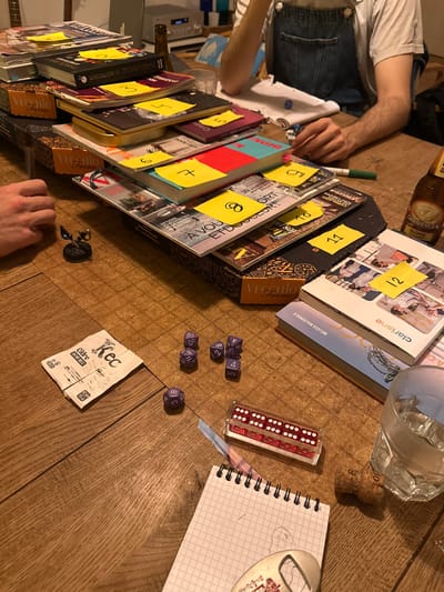 A table is set for a game. A series of books stacked at an angle has yellow sticky notes numbered 1-12. Next to them are several purple dice, a set of red dice in a clear case, and a notebook with sketches. A discarded cider label is visible. Parts of two people are in frame, one wearing overalls. A green marker, beer bottle, champagne cork, and a glass of water are also present on the wooden table.