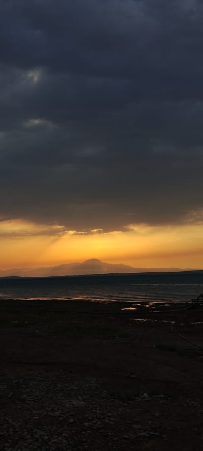 The image depicts a dramatic sunset over a body of water, likely Lake Chigdalik in Chigdalik, Tajikistan. Dark, brooding clouds dominate the upper portion of the frame, with shafts of golden light breaking through them. A range of mountains, silhouetted against the vibrant orange and yellow hues of the setting sun, form the horizon. The water is choppy, with ripples reflecting the sky's colors. The foreground consists of dark, rocky ground with patches of sparse grass. The scene evokes a sense of natural beauty and a touch of somberness due to the heavy clouds. No people or specific man-made structures are clearly visible, aside from what appears to be some industrial piping or tubing on the right side of the foreground.