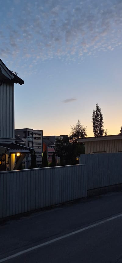 The image captures a view of a residential area at dusk in Lillehammer, Norway. In the foreground, a light-colored fence runs horizontally across the frame. Beyond the fence, several apartment buildings are visible, with some windows illuminated. Tall, dark trees are scattered between the buildings and in the mid-ground. The sky above is a gradient of soft blues and oranges, indicating the transition from day to night, with scattered clouds present. The overall scene is calm and residential, with no people or animals visible. The architecture suggests a Scandinavian setting.