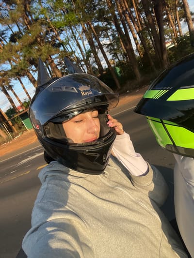 The image captures two individuals on what appear to be motorcycles or scooters, positioned on an asphalt road in the city of Mùi 3, Vietnam. In the foreground, a female subject is depicted from the upper torso, wearing a black full-face motorcycle helmet with a "Royal" crown logo and two dark, pointed ear/horn-like appendages attached to its top. Her clear visor is partially raised, revealing her face with eyes closed. Her right hand, exhibiting light pink nail polish, is touching the side of her helmet. She wears a light grey zippered hoodie over a light-colored long-sleeved garment. To her right, a second individual is partially visible, wearing a black and neon green full-face motorcycle helmet featuring geometric patterns, and light-colored clothing. The road beneath them displays a solid yellow line and a dashed white line. The background consists of a dense arrangement of tall, slender evergreen trees under bright daylight conditions, with a distant building and utility pole faintly visible through the foliage.
