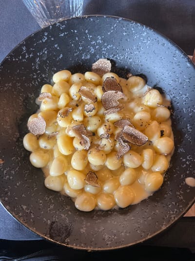 The image displays a close-up, top-down view of a single serving of gnocchi with truffle, positioned centrally within a dark, wide-rimmed ceramic bowl. The bowl exhibits a textured, speckled finish. The dish consists of numerous oval-shaped, pale yellow gnocchi coated in a creamy, light yellow sauce. Scattered across the gnocchi and sauce are multiple thin, irregularly shaped shavings of dark brown or black truffle, some displaying visible striations. Small black granular specks, consistent with ground pepper, are present on the food. In the upper left background, a portion of a clear, vertically ridged drinking glass is visible. The surrounding surface appears to be a dark, out-of-focus tablecloth, indicating a dining environment. This scene is located in a city within Valtournenche, Italy. No individuals are present in the frame.