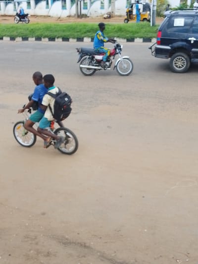 Two young boys ride a tandem bicycle on a dirt road. The boy in front wears a blue t-shirt and green shorts. The boy behind him wears a light-colored t-shirt and blue shorts and carries a black backpack. Behind them, a person in a blue and yellow outfit rides a red motorcycle. Further in the background, a black SUV is parked on the side of the road. A yellow motorized tricycle is also visible, with a person in a blue outfit standing next to it. The scene appears to be a street in Nigeria, possibly Oja Oba, Oko Erin, given the context. The sky is overcast, suggesting it might be a cloudy day. The overall impression is one of everyday life and transportation in a busy area.