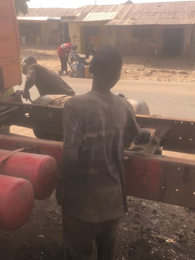 The image depicts an outdoor scene in a roadside mechanical repair area within the city of Tukur Tukur, Nigeria. The time is daytime, characterized by strong, hazy sunlight and a dusty atmosphere.
In the immediate foreground, a young male, observed from the rear, stands to the right of a large vehicle chassis. He is dressed in a heavily dirt-stained, light-colored short-sleeved t-shirt and dark pants. His head is turned slightly to the left, obscuring his facial features. His hands are not clearly visible, but he appears to be observing or engaged with the vehicle. The vehicle itself is primarily a truck chassis, featuring a prominent red frame and two red cylindrical air tanks mounted horizontally underneath, all covered in dust and grime. The ground directly beneath this area is loose, dark soil mixed with debris, consistent with a repair site.
To the left, another male individual, wearing a brown short-sleeved shirt and dark pants, is bent over the same vehicle's chassis. He is actively engaged in repair work, holding a tool in his right hand and interacting with the vehicle's undercarriage.
In the mid-background, across a dusty, unpaved road, a third individual is visible. This person wears a red shirt and is tending to a motorcycle or scooter. Nearby on the ground are several blue plastic jerry cans, commonly used for liquid storage, possibly fuel. Behind these figures, a row of single-story commercial buildings with corrugated metal roofs and light-colored, worn walls line the street, indicative of local businesses. The entire scene is permeated by a visible haze of dust.