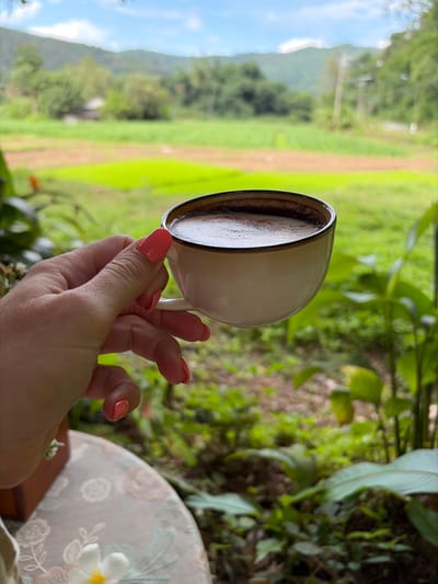 In Ban Huai Takhrai, Thailand, a hand with coral-colored fingernails holds a white coffee cup filled with dark coffee. The cup is positioned in the foreground, with a blurred background of lush green fields and distant mountains under a cloudy sky. The table surface beneath the cup is decorated with a floral pattern.