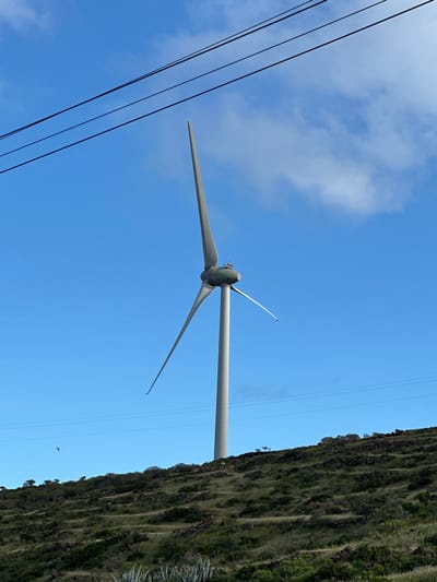 A three-bladed wind turbine is positioned on a verdant, terraced hillside in San Sebastián de la Gomera, Spain. The turbine features a light gray cylindrical tower topped by a dark gray nacelle. Its three blades are stationary: one extends vertically upwards, another diagonally downwards to the left, and the third diagonally downwards to the right. The hillside exhibits sparse green and brown scrub vegetation interspersed with dark exposed rocks. Above the turbine, against a bright blue sky with scattered white clouds, three parallel dark power lines traverse the upper left quadrant of the image. A thinner, dark horizontal wire runs across the middle ground, below the turbine's nacelle. A small dark bird is visible in flight in the mid-left sky. No individuals are present.
