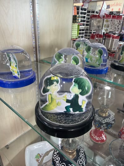 This is a medium shot, eye-level, slightly angled, indoor photograph of decorative items on store shelves in Acıpayam, Turkey. The foreground displays multiple clear plastic domes with blue and black bases, each containing a scene of cartoon figures. One dome features a green-haired character in a yellow dress with a crown, and another shows two green-haired figures holding hands. Behind these, other similar domes are visible. The background includes shelves filled with various packaged goods, including what appear to be craft or hobby supplies, and signage in Turkish. A string of LED lights runs vertically along the left side of the frame, illuminating the display. The overall impression is of a retail environment selling novelty items.