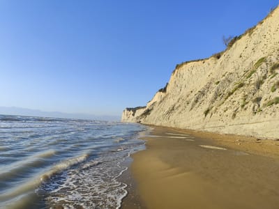 The image displays a coastal landscape in Agios Stefanos, Greece, under a clear, light blue sky. On the right, a prominent, high cliff of light beige, stratified rock extends diagonally from the upper right toward the mid-left horizon. The cliff face exhibits vertical erosion patterns and horizontal layering, with scattered green vegetation and a few dry, leafless trees on its upper edge and slopes. A wide, sandy beach, colored golden-brown, occupies the foreground and mid-ground, sloping gently towards the sea on the left. The sand shows ripples and variations in wetness, with minimal driftwood debris visible near the cliff base. The sea on the left features multiple parallel wave fronts, with white foam from breaking waves receding onto the shore. The water appears a muted greenish-blue. In the far distance, faint, hazy blue mountains are discernible on the horizon. No people or man-made objects are present in the scene.