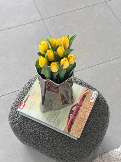 The scene is an indoor environment located in Tielrode, Belgium. A metallic, irregularly shaped, silver-toned vase containing approximately 10-12 yellow tulips is positioned centrally. The tulips feature green stems and leaves, with blossoms mostly closed or partially open. The vase rests upon a magazine titled "AUTO Classiccars" with "PORSCHE" prominently displayed, featuring a red Porsche sports car on its cover. This magazine, in turn, is placed on a dark gray, shaggy-textured, cylindrical or oval stool. The background consists of a light gray tiled floor with visible grout lines. A section of a lighter-colored, shaggy rug is visible on the lower right, adjacent to the stool. No people or animals are present.