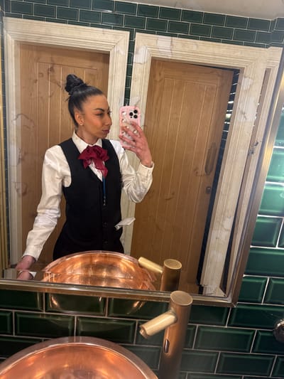A person is taking a selfie in a bathroom. The person is wearing a white shirt with a black vest and a red bow tie. They are holding a pink phone with a Hello Kitty case. The bathroom has green tile walls and a wooden door. There are copper-colored sinks. The location is Warrington, United Kingdom.