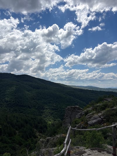 The image depicts a panoramic view from a rocky, elevated vantage point overlooking a lush, forested valley. The sky above is a vibrant blue, adorned with scattered, fluffy white clouds. The foreground features a rustic wooden railing, weathered and aged, with a few exposed rocks. Beyond the railing, the terrain drops into a dense, dark green forest that covers the slopes of a large hill or mountain. In the distance, the valley opens up to reveal more rolling hills and a distant settlement with scattered buildings, suggesting a rural area. The overall impression is one of natural beauty and tranquility. The location is identified as being in Dolno Drianovo, Bulgaria. There are no people or animals visible in the image.