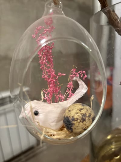 A decorative glass orb contains a small, white artificial bird nestled in straw next to a speckled brown and beige egg. A sprig of bright pink dried flowers extends upwards from behind the bird. The orb is suspended and appears to be part of a display. In the background, a cylindrical glass container holding a dried branch is visible, along with a concrete-like wall. The scene is indoors, likely in a shop or home. The location is identified as Novaya Usman, Russia.