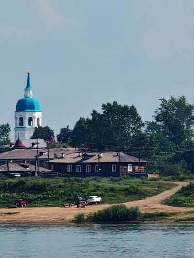 In Yeniseysk, Russia, this image captures a serene waterscape with a view of the cityscape. In the foreground, a body of water reflects the light. The middle ground features a sandy bank with several vehicles, including a red car, a boat trailer, and a white car, suggesting a recreational or transport area. A dirt road winds through the grassy embankment behind the vehicles. The background reveals a collection of wooden houses and a distinctive white church with a blue dome and steeple, a prominent landmark. Lush green trees dot the landscape, contributing to the overall natural beauty of the scene. The sky above is a clear, light blue.