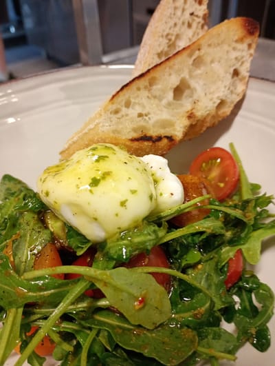 A close-up shot captures a plate of food, likely a salad with a poached egg, served on a white plate with a subtle brown rim. Two slices of toasted bread, with a golden-brown crust and a soft, airy interior, are placed atop the salad. The salad itself consists of vibrant green arugula, interspersed with halved cherry tomatoes and what appears to be a drizzle of dressing. A poached egg, glistening with dressing and flecked with herbs, sits prominently on the salad. The background is blurred, suggesting an indoor setting, possibly a restaurant, with hints of metallic surfaces and muted colors. The image does not depict any specific location in the city of Tingstäde, Sweden.