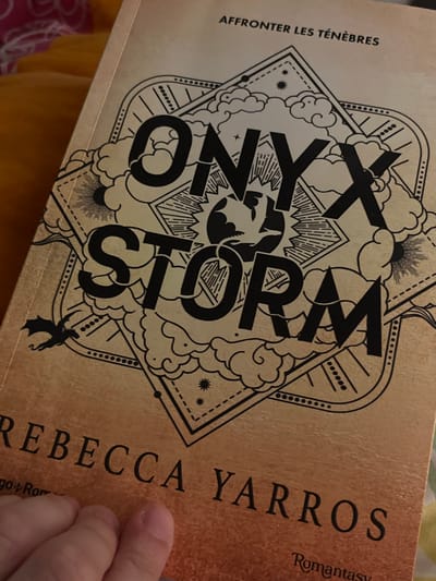A book cover with a gold background features a large, diamond-shaped design. Inside the diamond, bold black lettering spells "ONYX STORM." Below the title is a stylized drawing of a bat silhouette. The diamond is adorned with clouds and stars. The book's author is listed on the right side of the cover as REBECCA YARROS in black capital letters. "Romantasy" is printed as a description near the bottom. The title "affronter les ténèbres" is printed on the side.